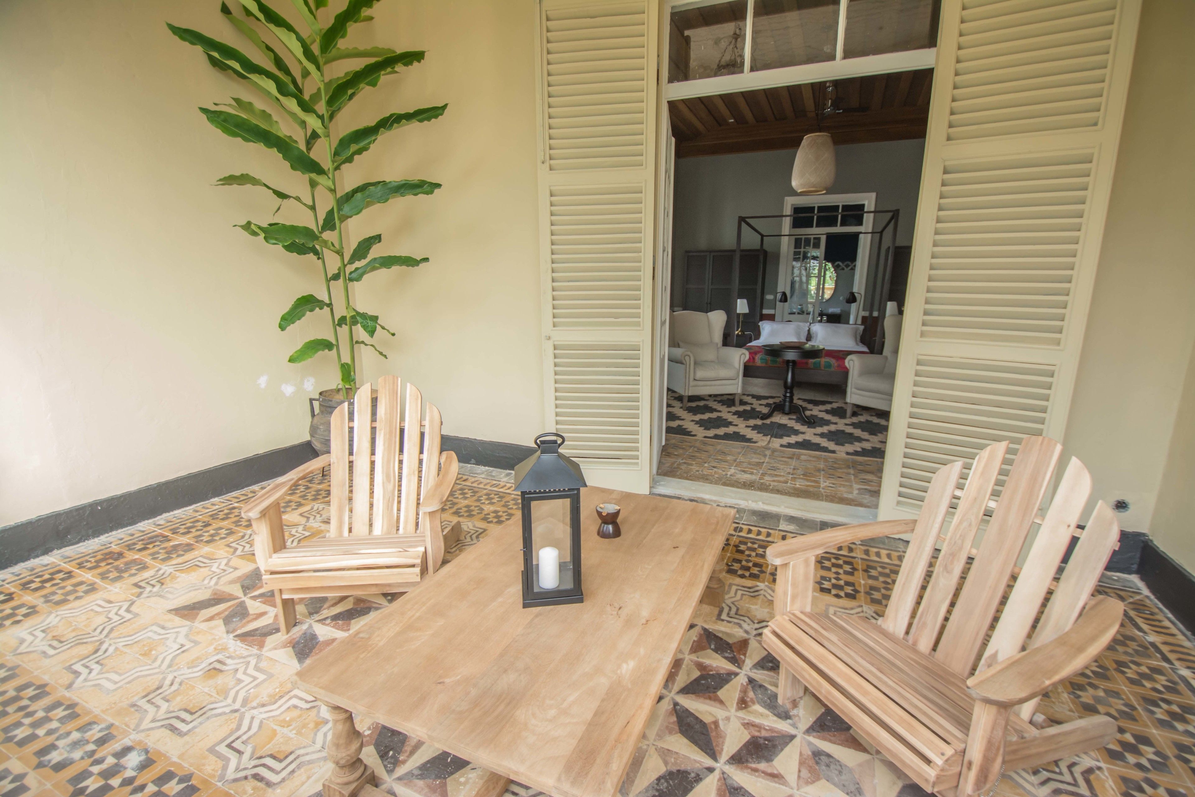 A wooden table with chairs, a lantern, and a potted plant in a room with patterned floor tiles and wooden shutters.