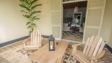 A wooden table with chairs, a lantern, and a potted plant in a room with patterned floor tiles and wooden shutters.