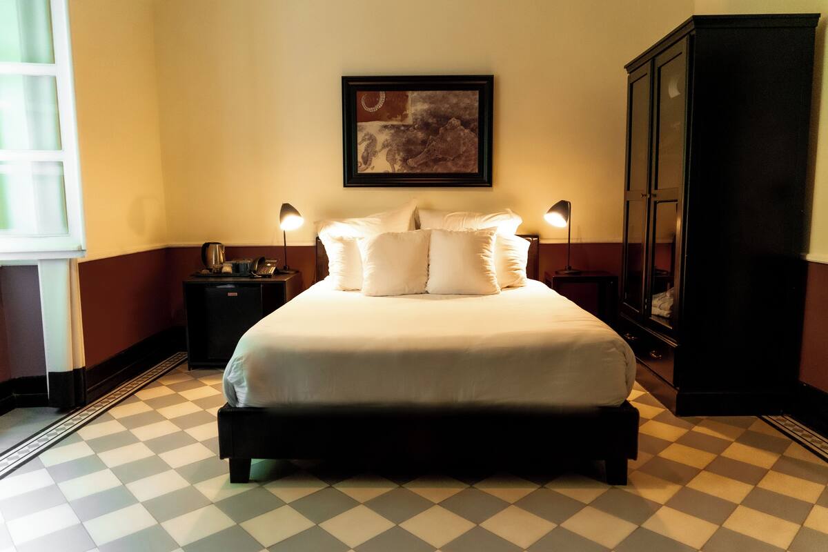 A neatly made bed with white linens and pillows, a framed artwork above it, and a checkered floor pattern.