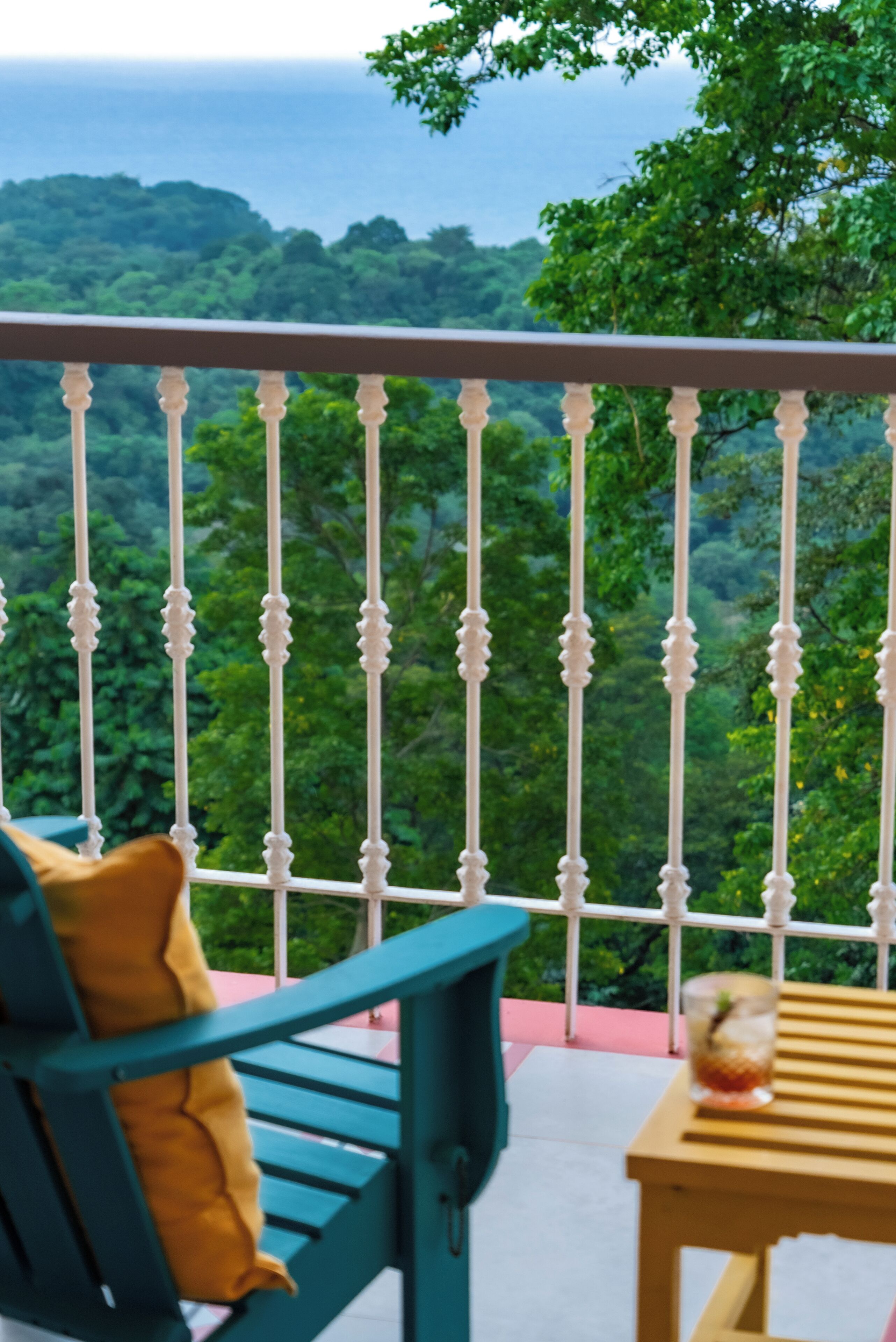 A balcony with a white balustrade, a teal chair with a yellow cushion, and a wooden table with a glass.