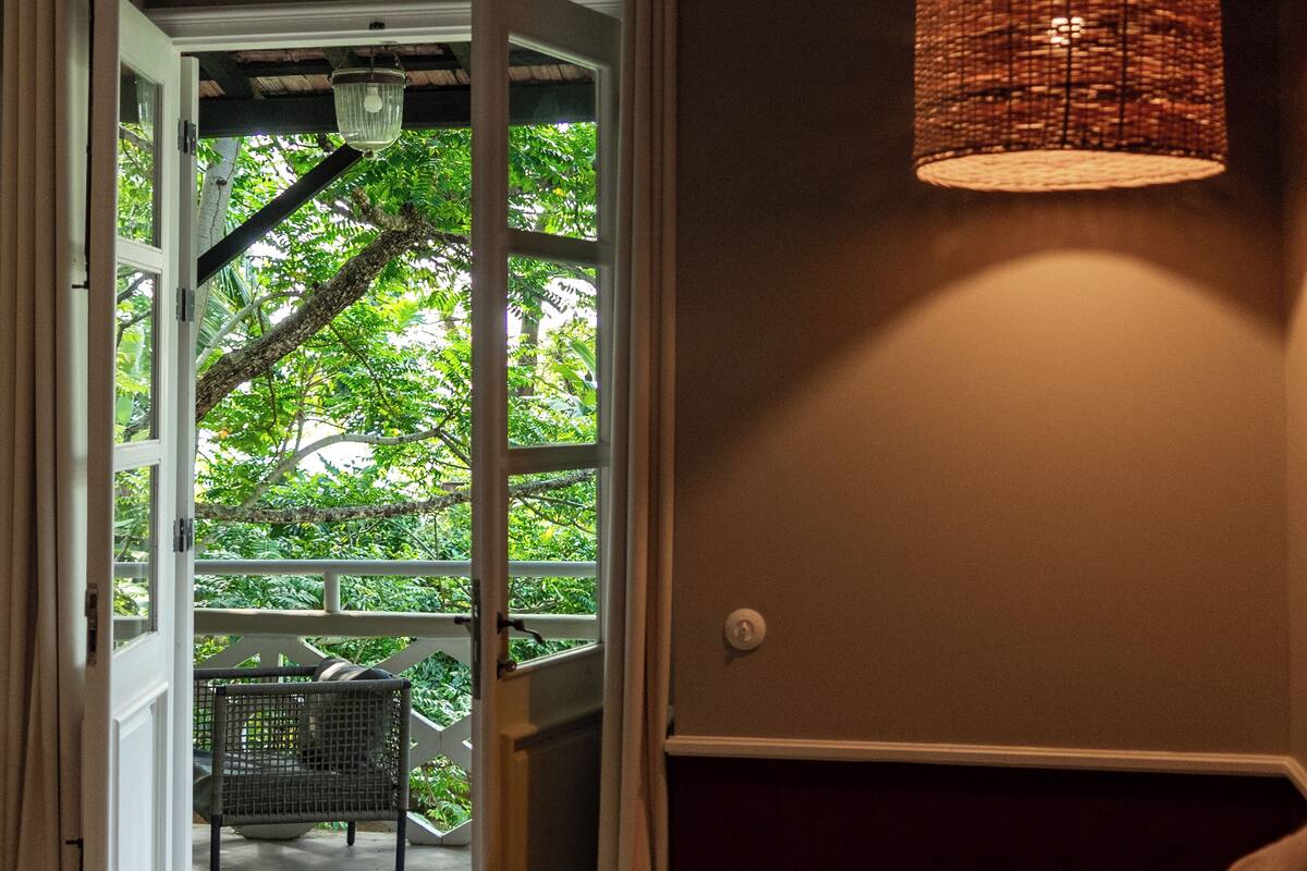 An open doorway leading to a balcony with a wicker lamp, a chair, and a view of greenery.