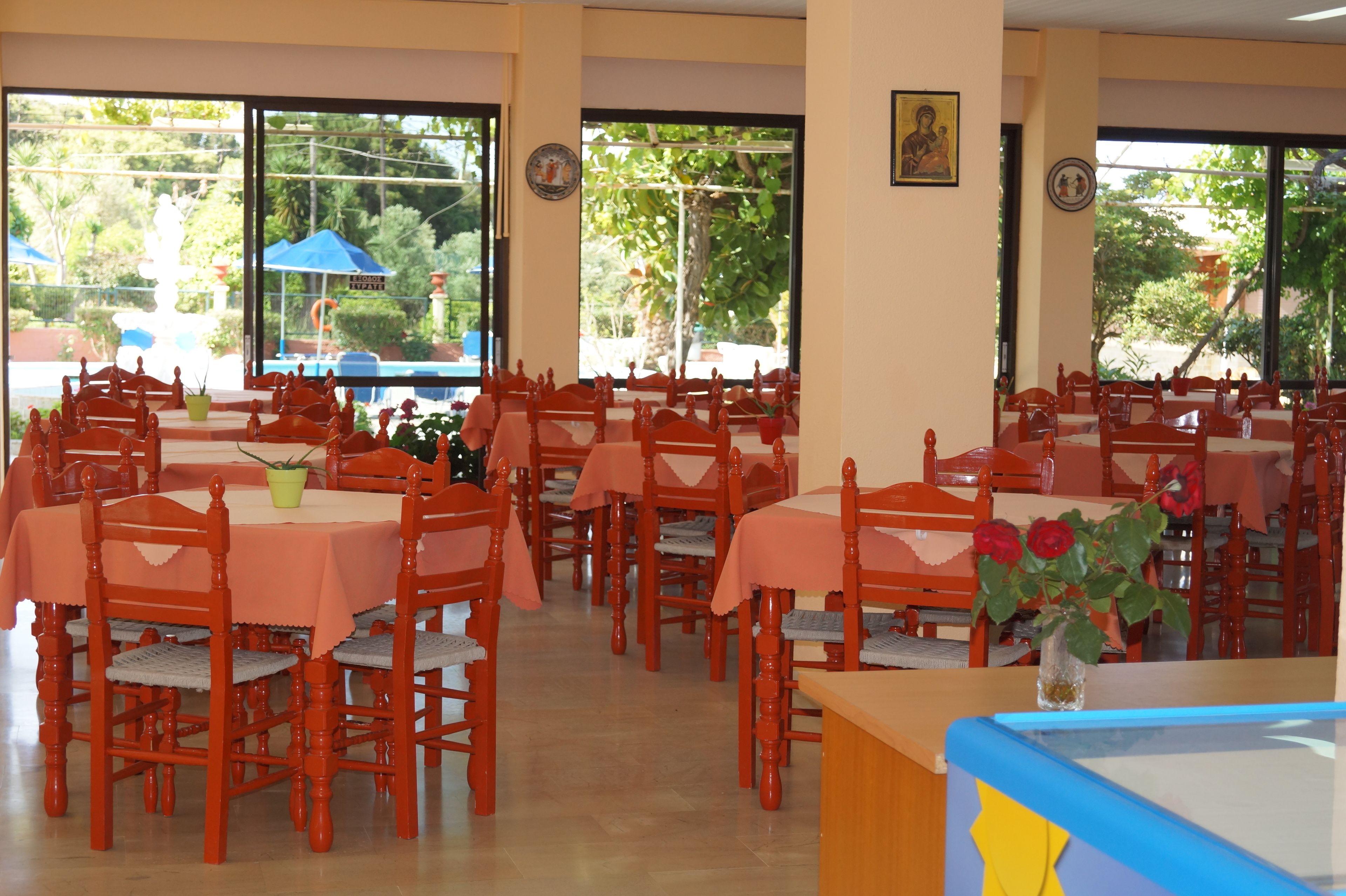 A dining area with multiple tables and chairs, large windows with a view of greenery outside, and a few potted plants.