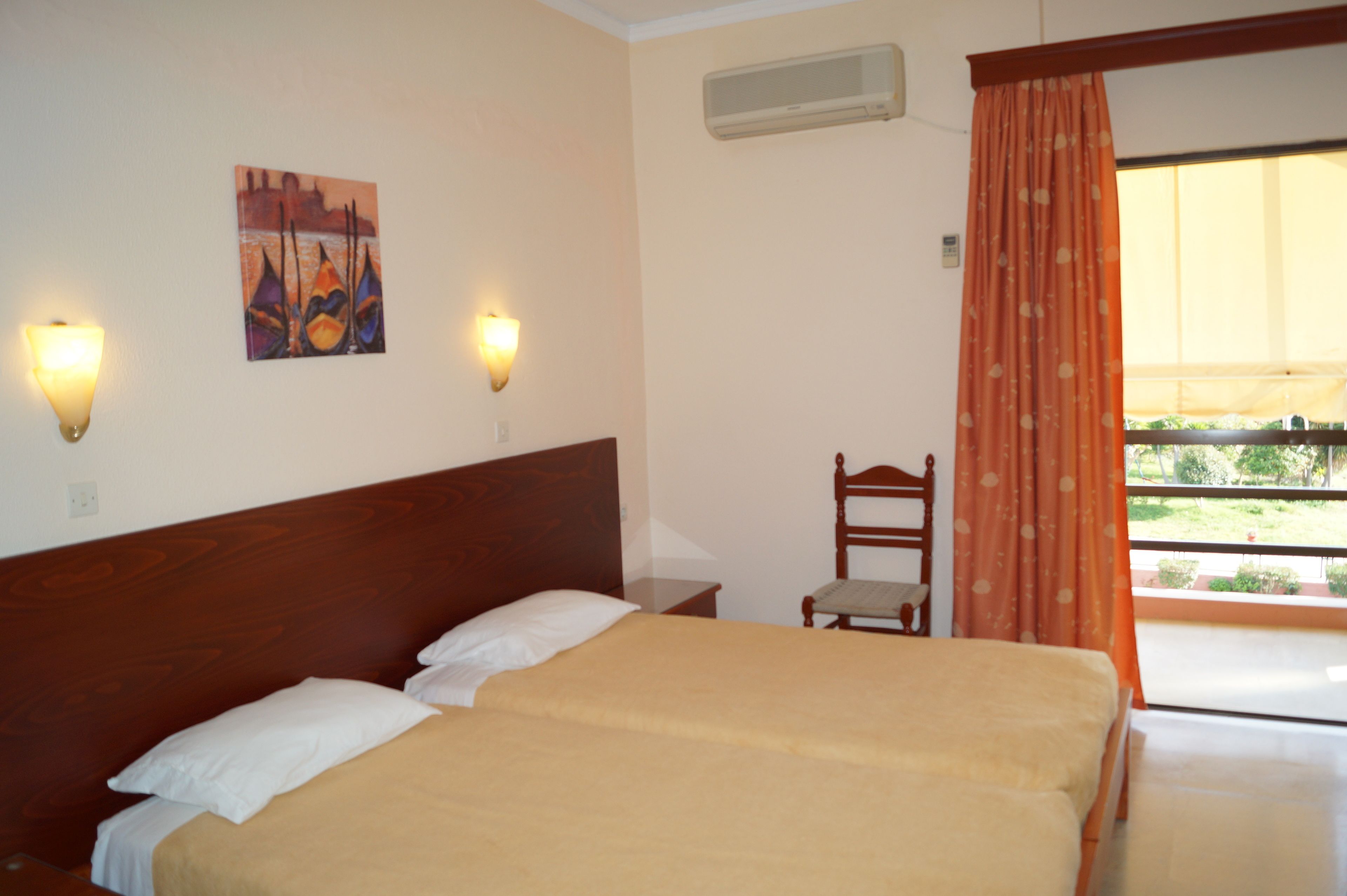 A double bed with a wooden headboard, two bedside lamps, a small table, a chair, and a painting on the wall.