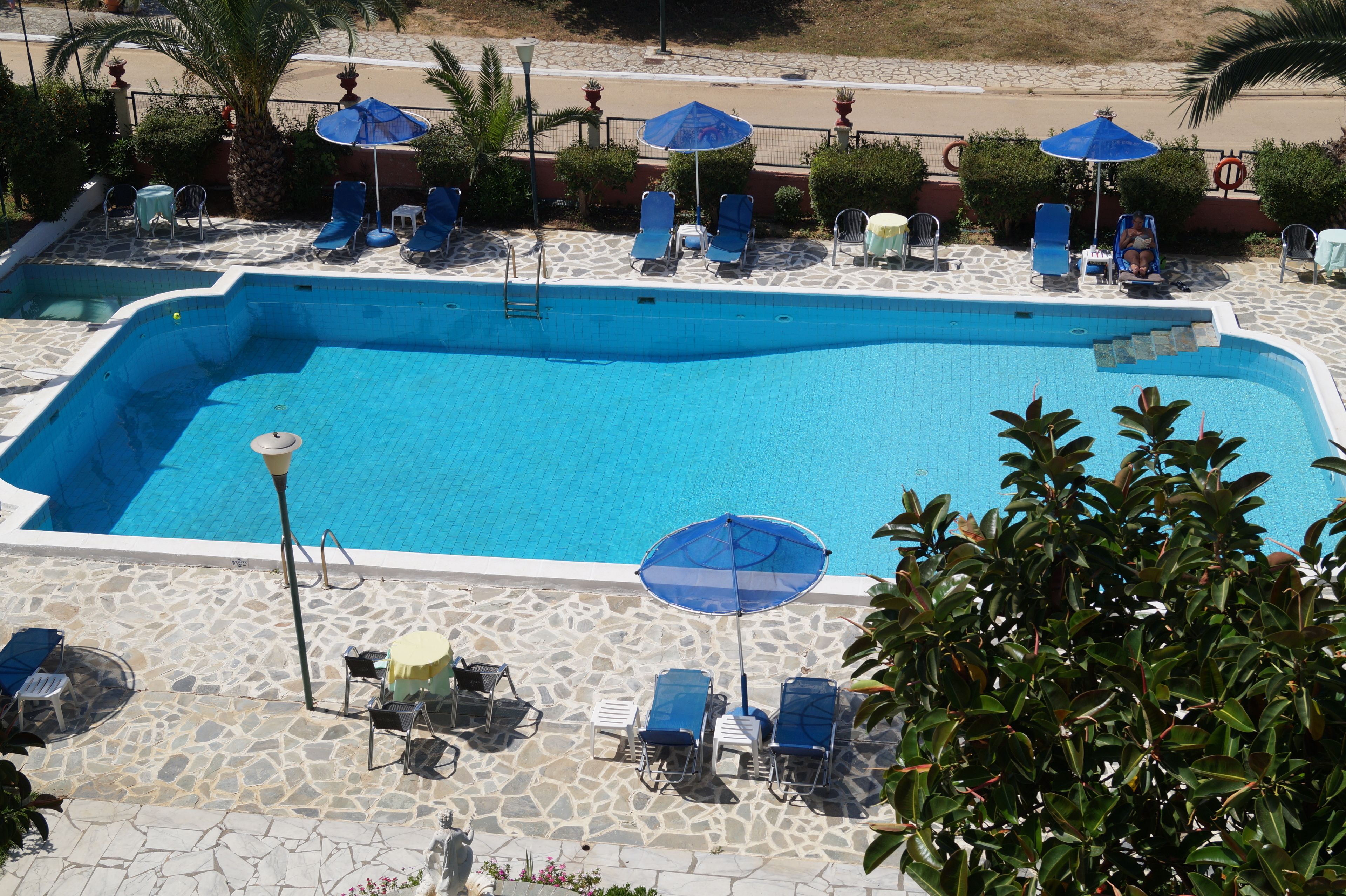 Piscina con sillas de descanso y sombrillas, rodeada de baldosas de piedra y follaje.