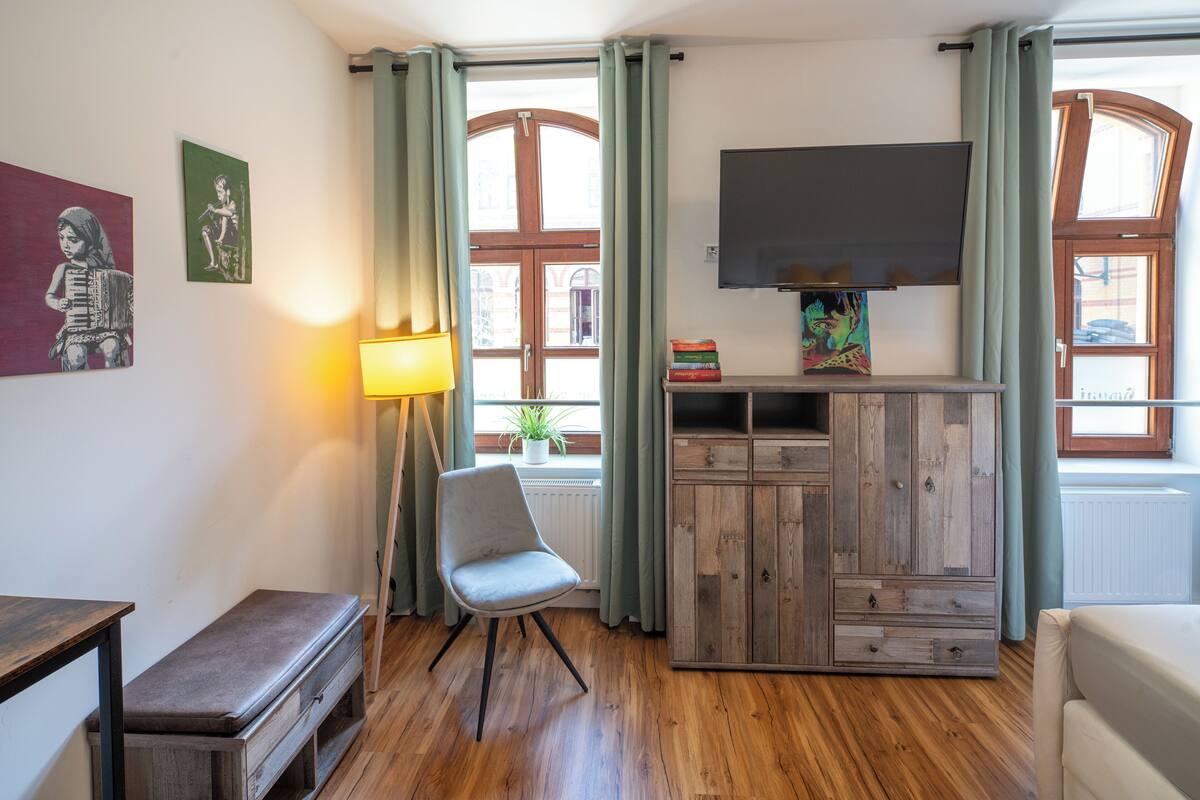 A room with a wooden floor, a bench, a chair, a TV mounted on the wall, and a window with curtains.
