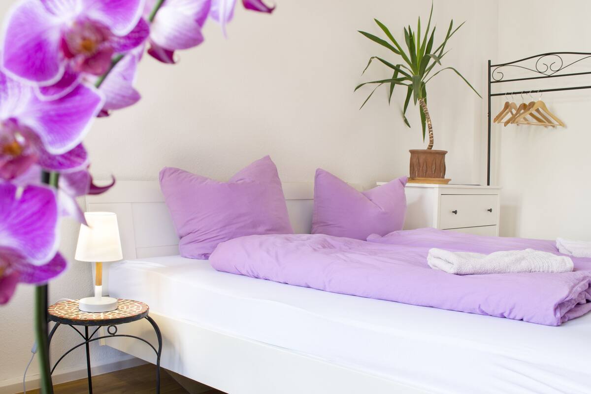 A neatly made bed with purple bedding, a bedside table with a lamp, a potted plant, and a metal bed frame.