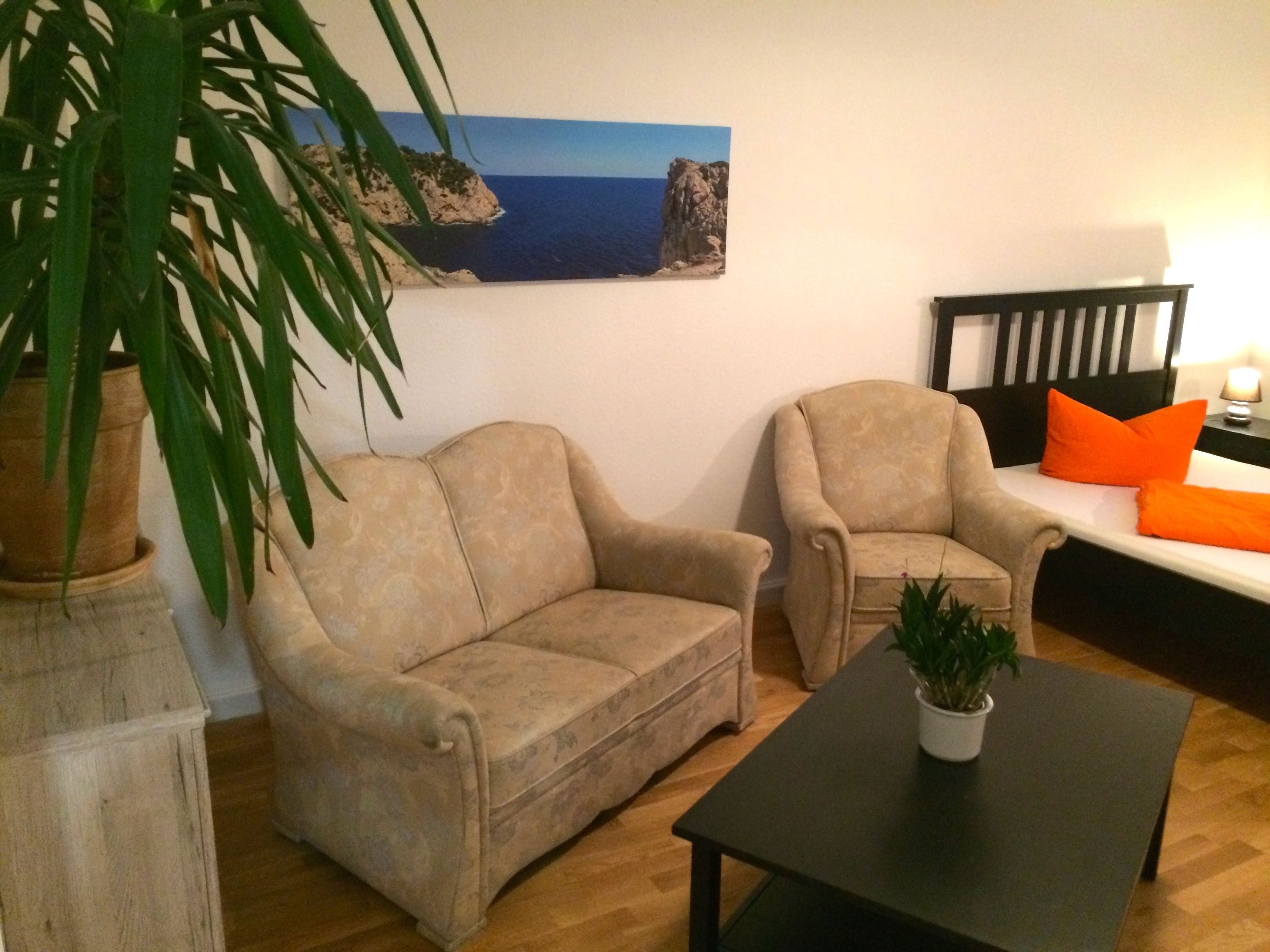 Ein Wohnzimmer mit einem beigen Sofa, einem passenden Sessel, einem kleinen Tisch mit Pflanze und einem Gemälde einer Küstenlandschaft an der Wand.