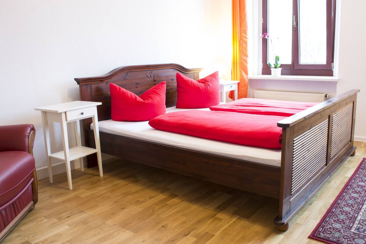 A wooden bed with red bedding, a nightstand, a red chair, and a window with curtains.