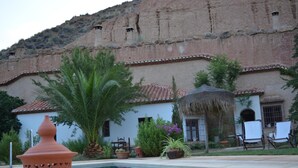 Zona de piscina con un edificio blanco al fondo, rodeada de vegetación y una montaña.