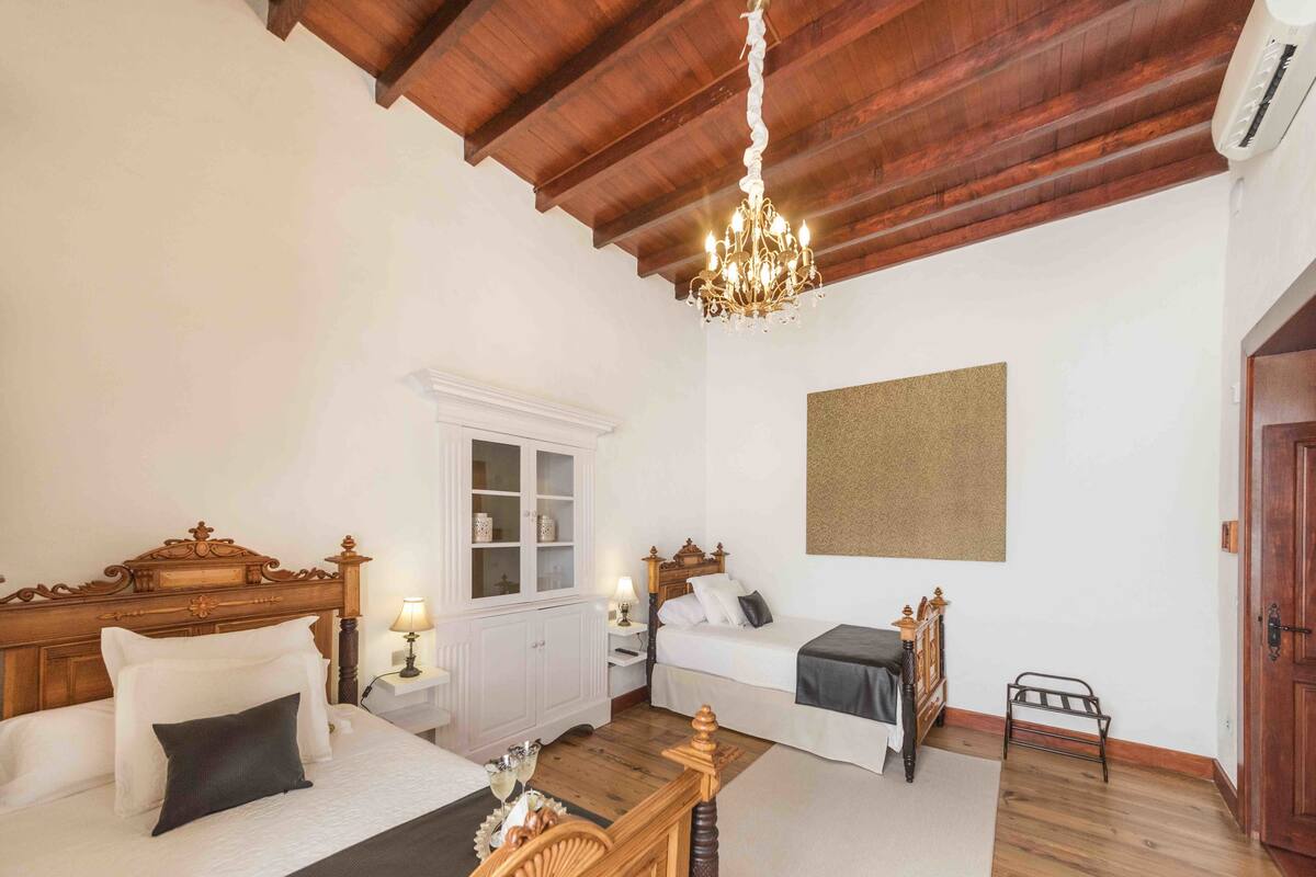 A room with a wooden ceiling, a chandelier, a bed with a wooden headboard, a bedside table with a lamp, a small cabinet, and a wall-mounted artwork.