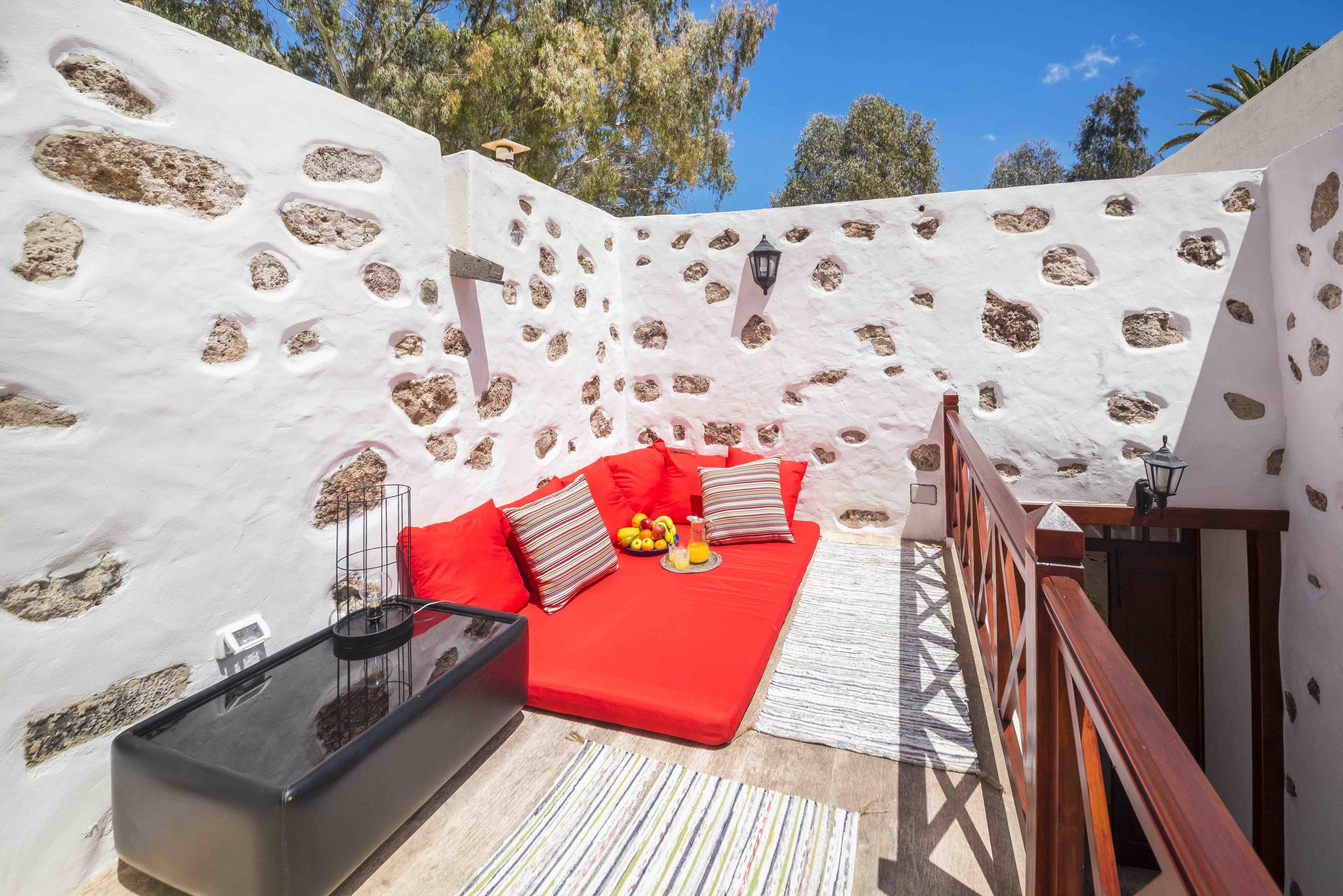 A cozy outdoor seating area with a red sofa, cushions, and a small table with fruits and a drink.