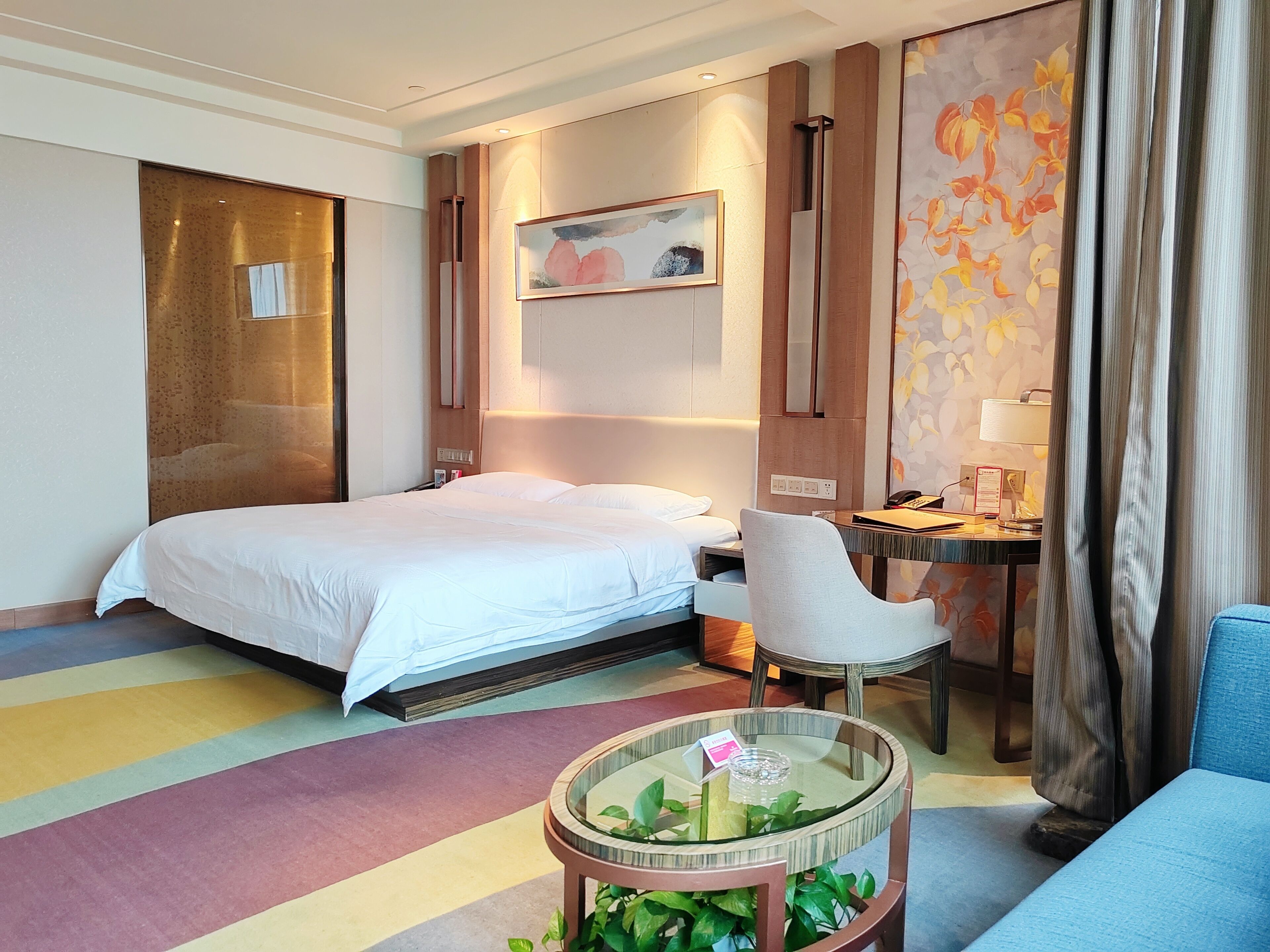 A hotel room with a bed, a desk, a chair, and a small table with a plant.
