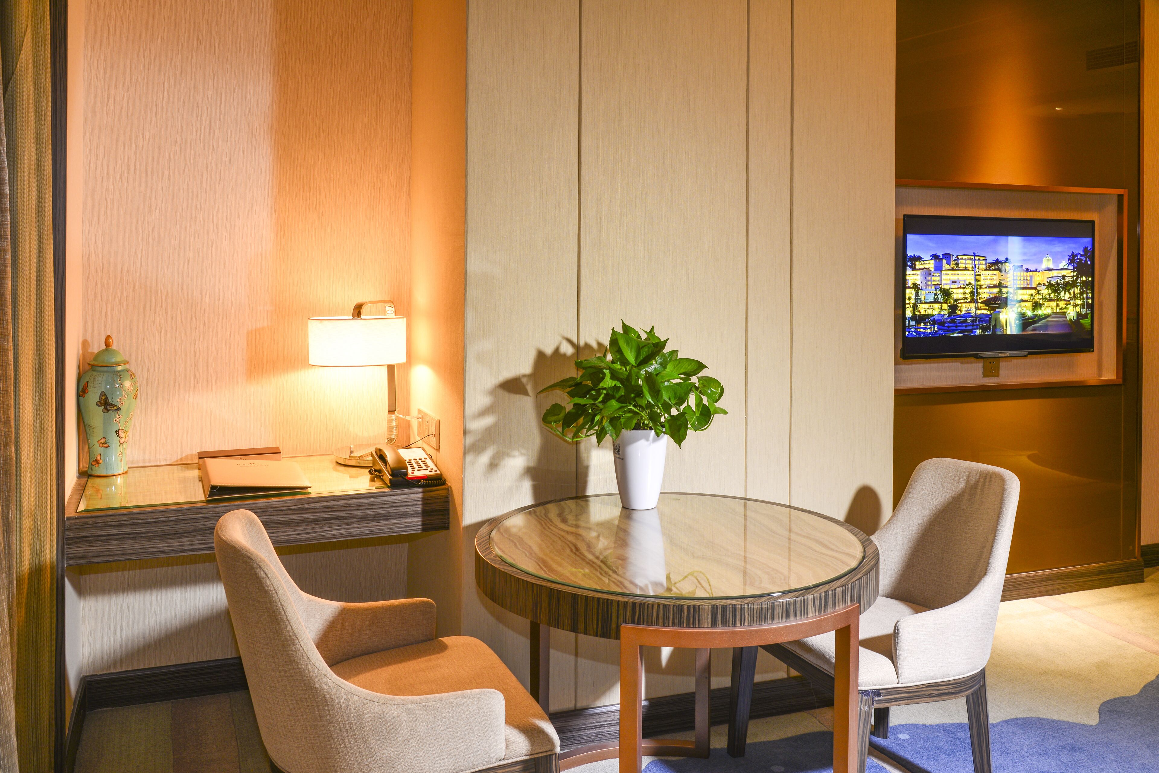 Ein Hotelzimmer mit einem runden Glastisch, zwei Stühlen, einer Topfpflanze, einer Lampe, einem Fernseher und einer dekorativen Vase.
