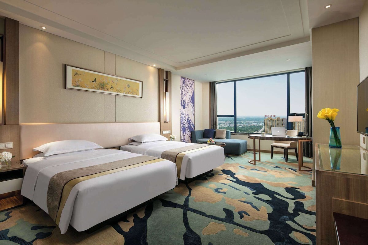 Habitación de hotel con dos camas, un escritorio, una silla, un ventanal y vistas a la ciudad.
