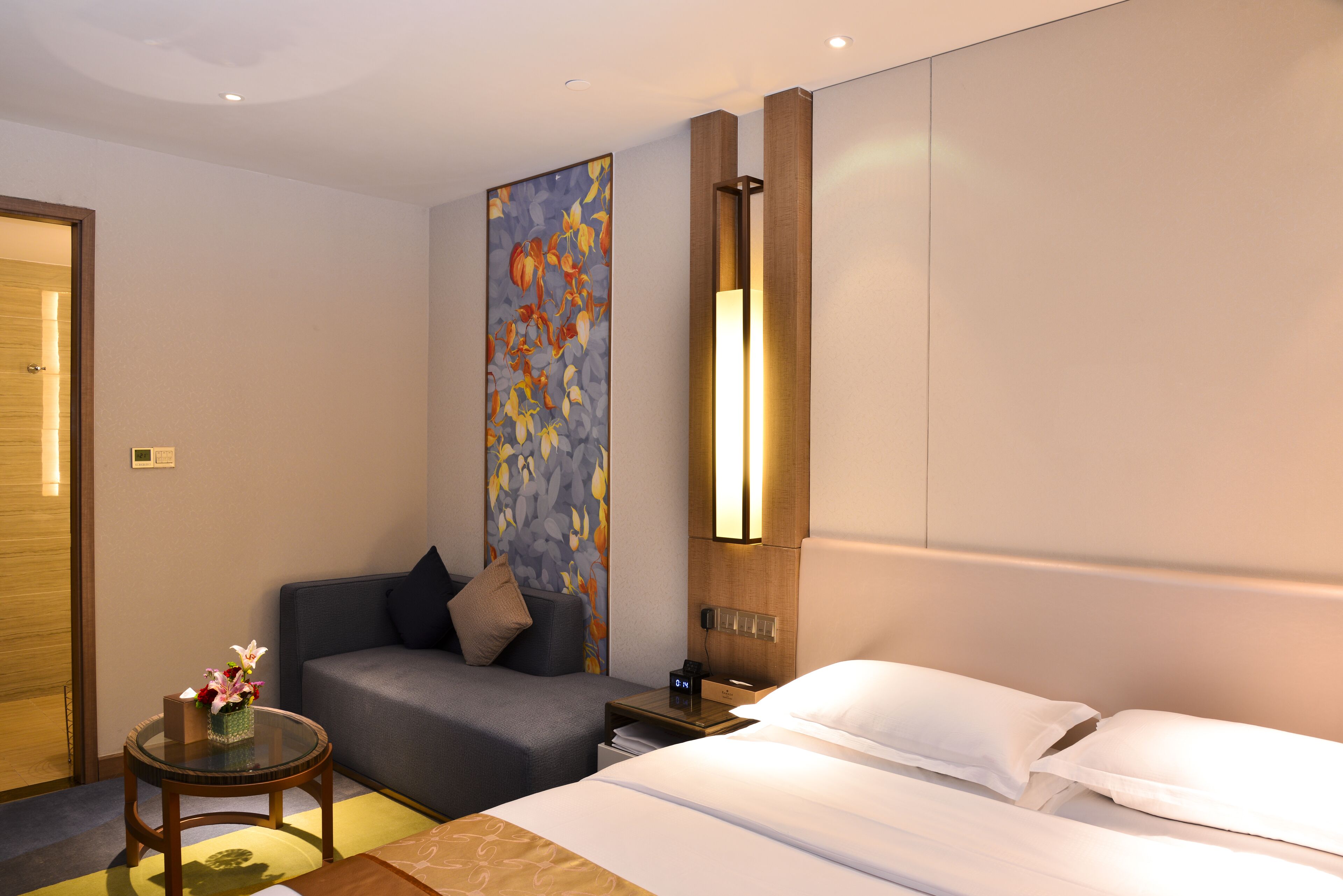 A hotel room with a bed, a sofa, a small table with a vase of flowers, a nightstand with a lamp, and a wall with a colorful artwork.