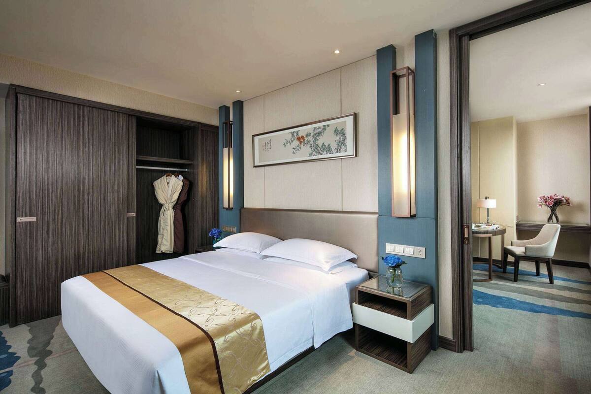 Una habitación de hotel con una cama grande, un armario, una mesita de noche con un florero y una silla.