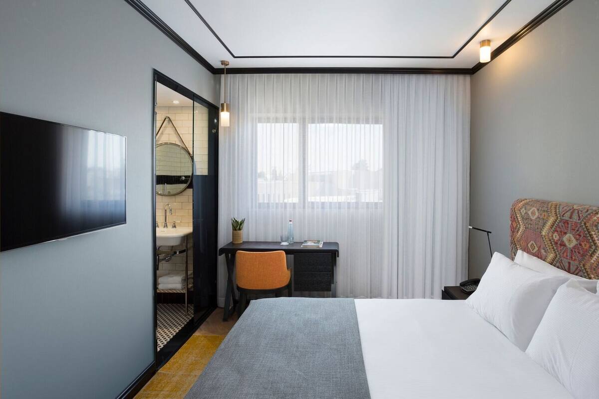 A hotel room with a bed, a desk with a chair, a TV, a window with curtains, and a patterned headboard.