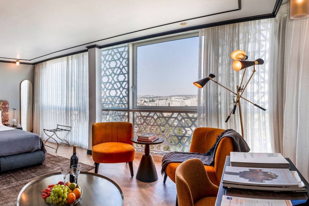 A modern hotel room with a large bed, a sitting area with a coffee table, and a view of the city through large windows.