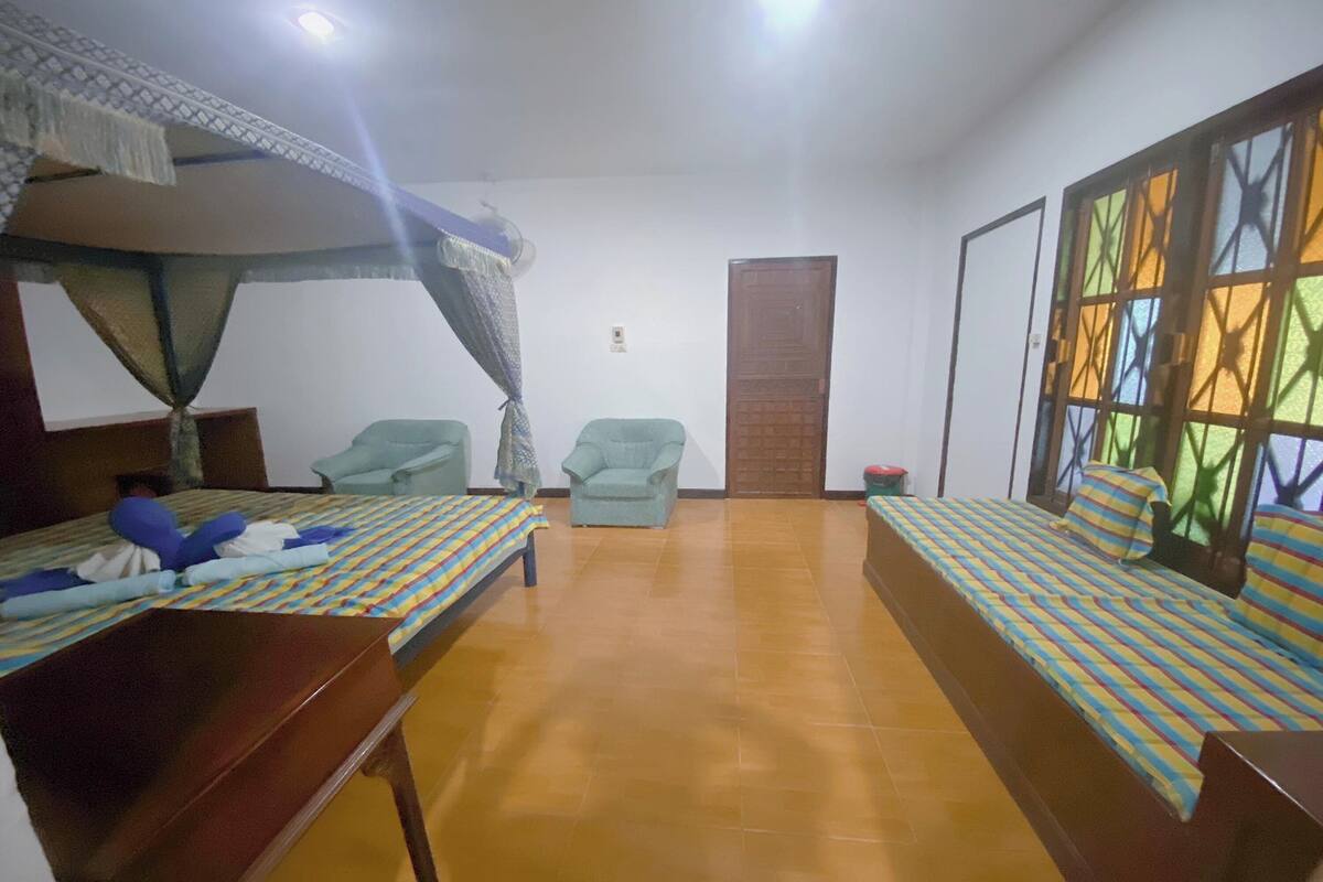 A room with two beds, a bench, two chairs, a wooden door, and a window with a geometric pattern.