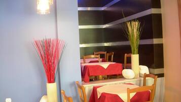 A dining area with tables set for four, each with a red tablecloth. There are wooden chairs and a vase with red and white decorative elements.