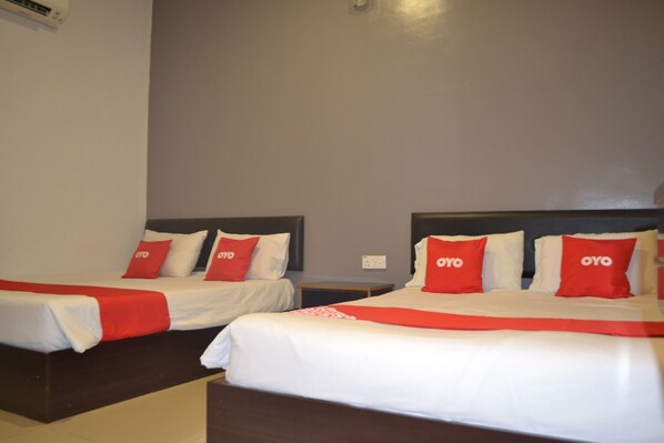 Two single beds with white bedding and red pillows, each with the OYO logo, are positioned against a neutral wall, accompanied by a bedside table.