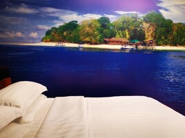 Una cama con ropa blanca, vista a una playa con palmeras y una cabaña pequeña junto al agua.