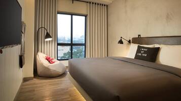 A modern hotel room with a large bed, a bean bag chair, a window with curtains, and a city view.