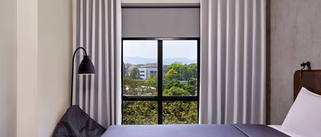 Habitación de hotel con un ventanal grande, una cama, una lámpara de pie negra y vistas a zonas verdes y edificios.