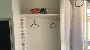 A small, narrow room with a white brick wall, a white door, and a white refrigerator. There are two black hangers on a white shelf, and a window with white curtains. - Pousada Aldeia Buzios (Búzios)