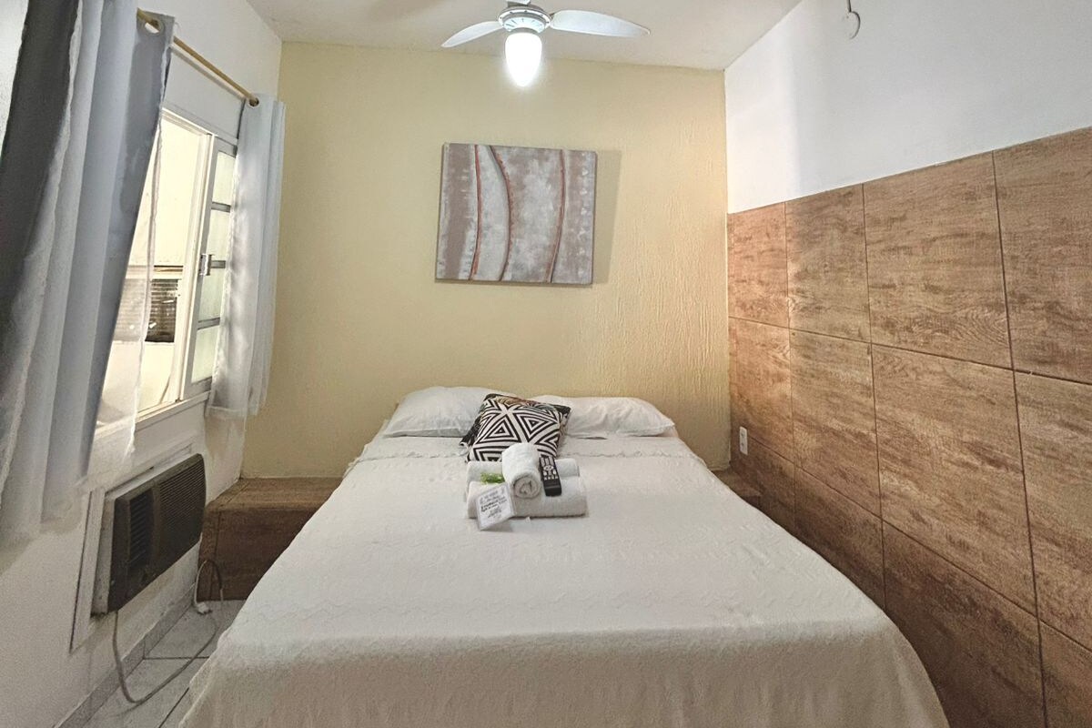 A hotel room with a bed, a ceiling fan, a window with curtains, and a wall-mounted artwork.