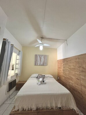A hotel room with a bed, a ceiling fan, a window with curtains, and a wall-mounted artwork.
