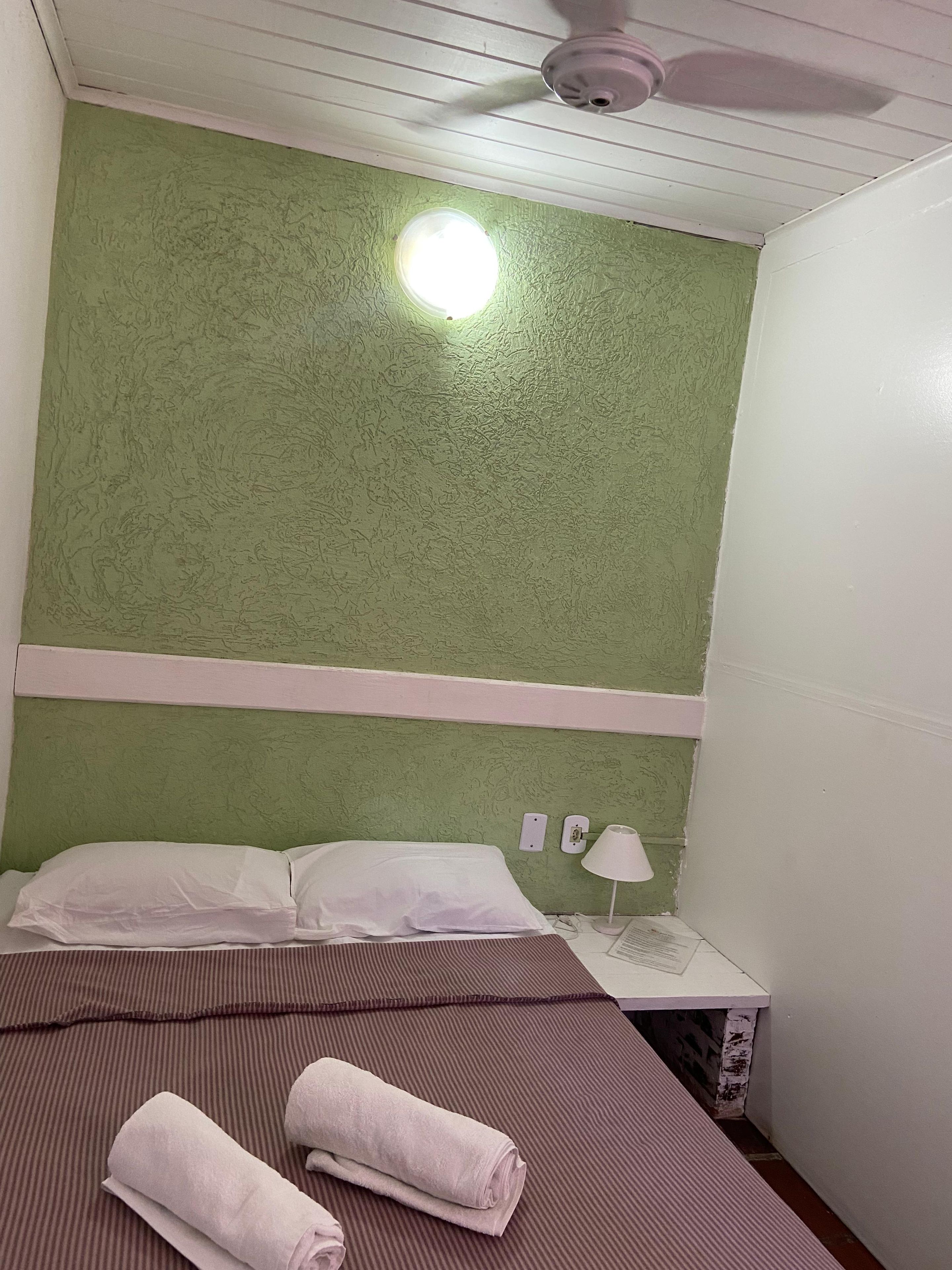 A room with a green textured wall, a ceiling fan, a bed with white pillows and a striped brown bedspread, and a small bedside table with a lamp.