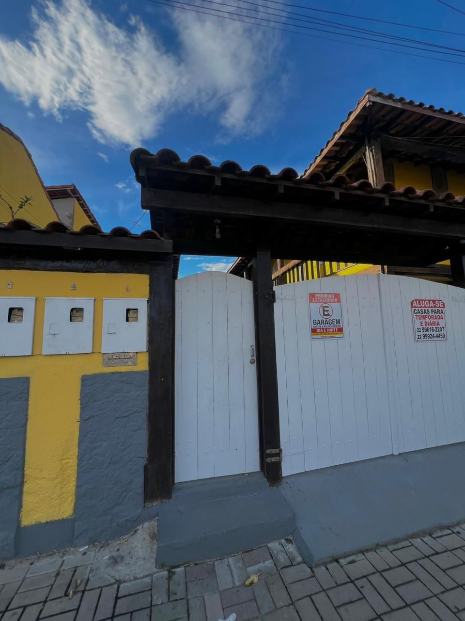 A gated entrance with a wooden pergola, a white fence, and a yellow building with a blue door.