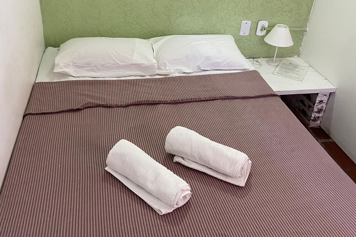 A small, single-bed room with a green textured wall, a white bedside table with a lamp, and two rolled towels on the bed.
