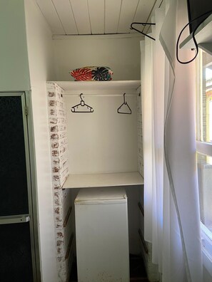 A small, narrow room with a white brick wall, a white door, and a white refrigerator. There are two black hangers on a white shelf, and a window with white curtains. - Pousada Aldeia Buzios (Búzios)
