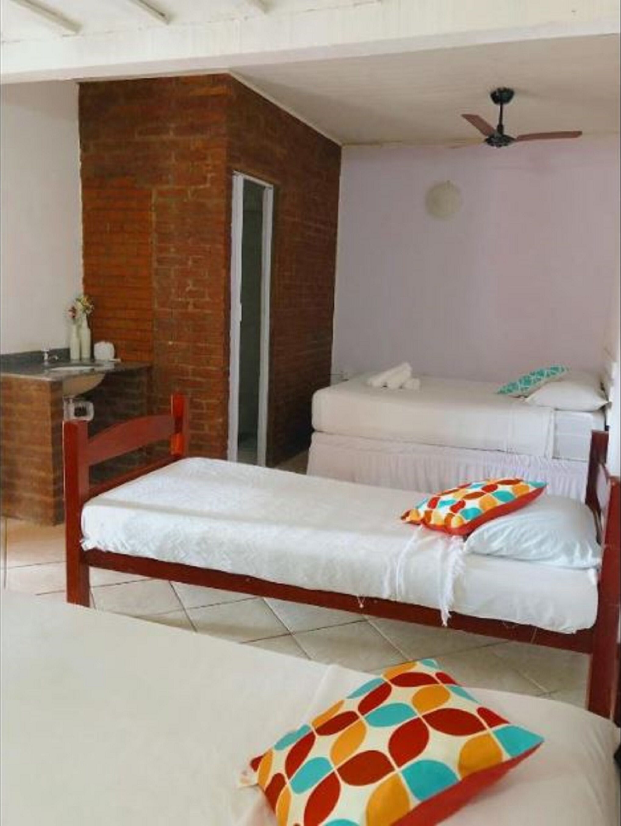 A single bed with a colorful pillow, a wooden headboard, and a ceiling fan in a room with a brick wall and a white door.