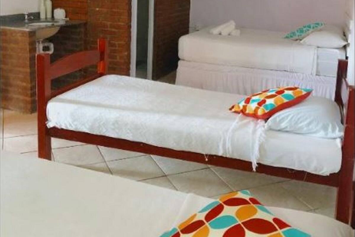 A single bed with a colorful pillow, a wooden headboard, and a ceiling fan in a room with a brick wall and a white door.