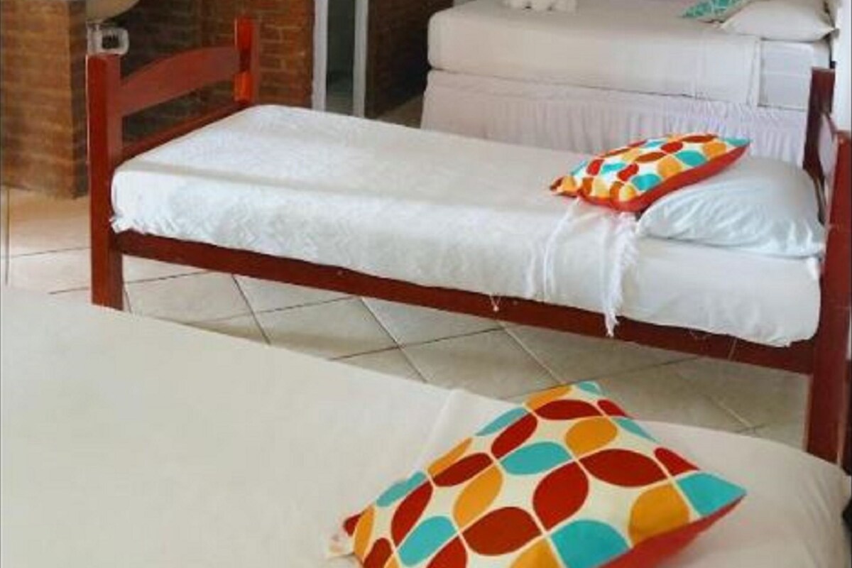 A single bed with a colorful pillow, a wooden headboard, and a ceiling fan in a room with a brick wall and a white door.