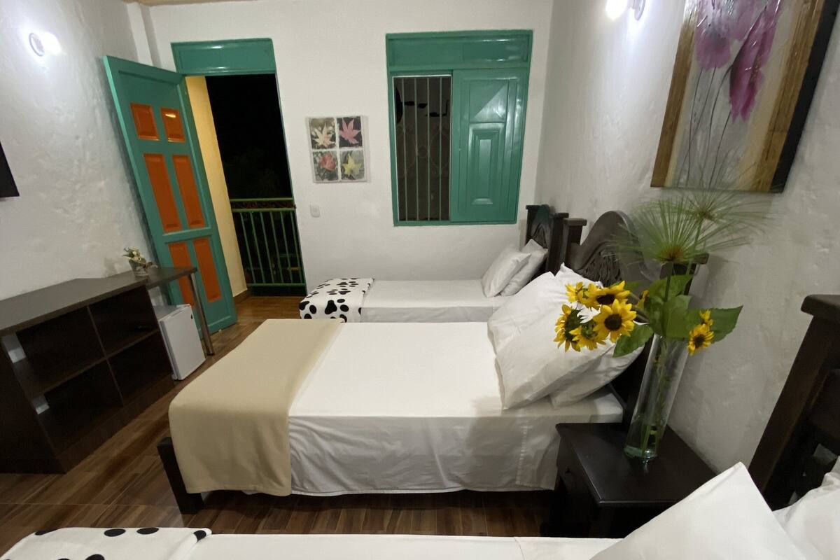 Une chambre à coucher avec un lit, une table de chevet, un vase de fleurs et une fenêtre avec des volets verts.