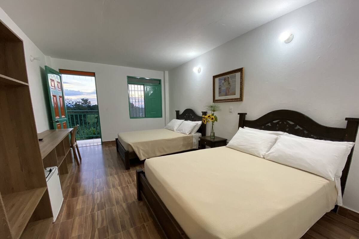 Une chambre avec deux lits, un bureau en bois, une chaise, une petite table avec un vase et une porte donnant sur un balcon.