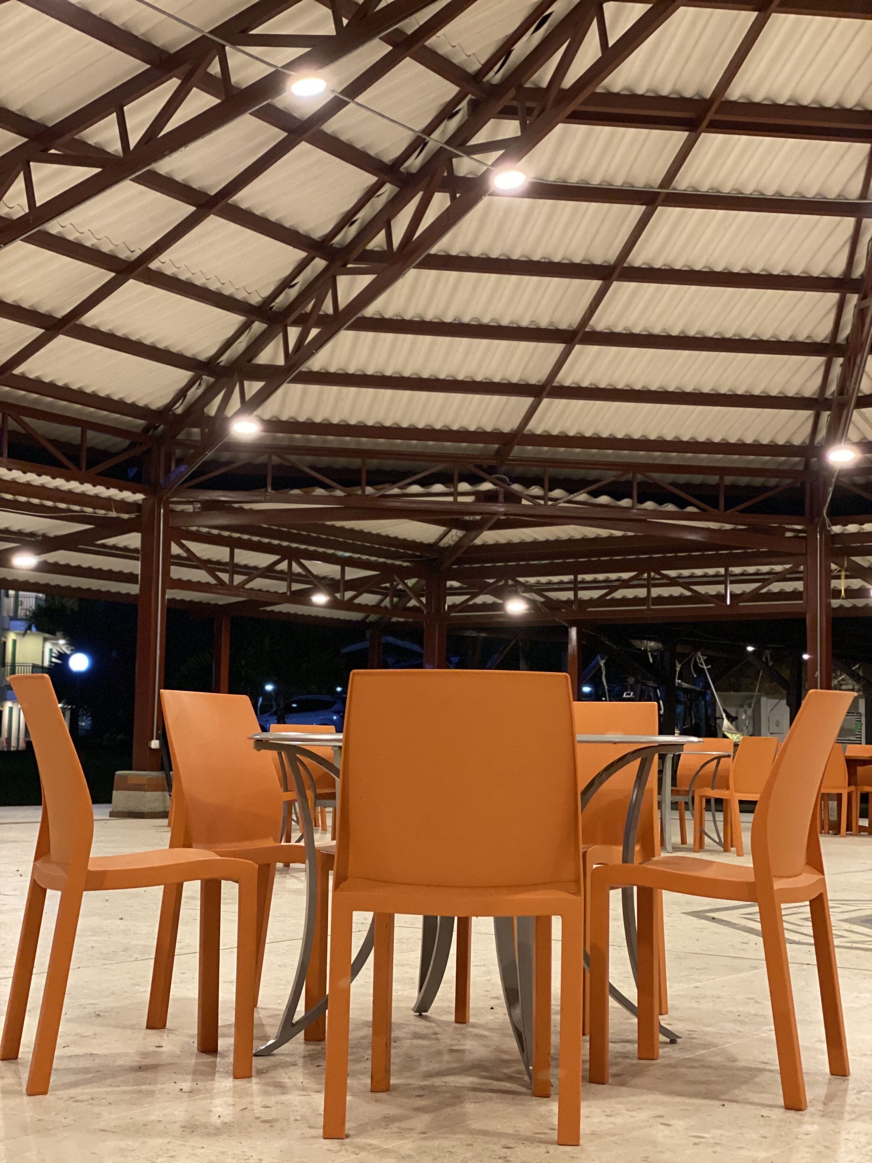 A covered outdoor seating area with orange chairs and tables, metal beams, and a corrugated metal roof.