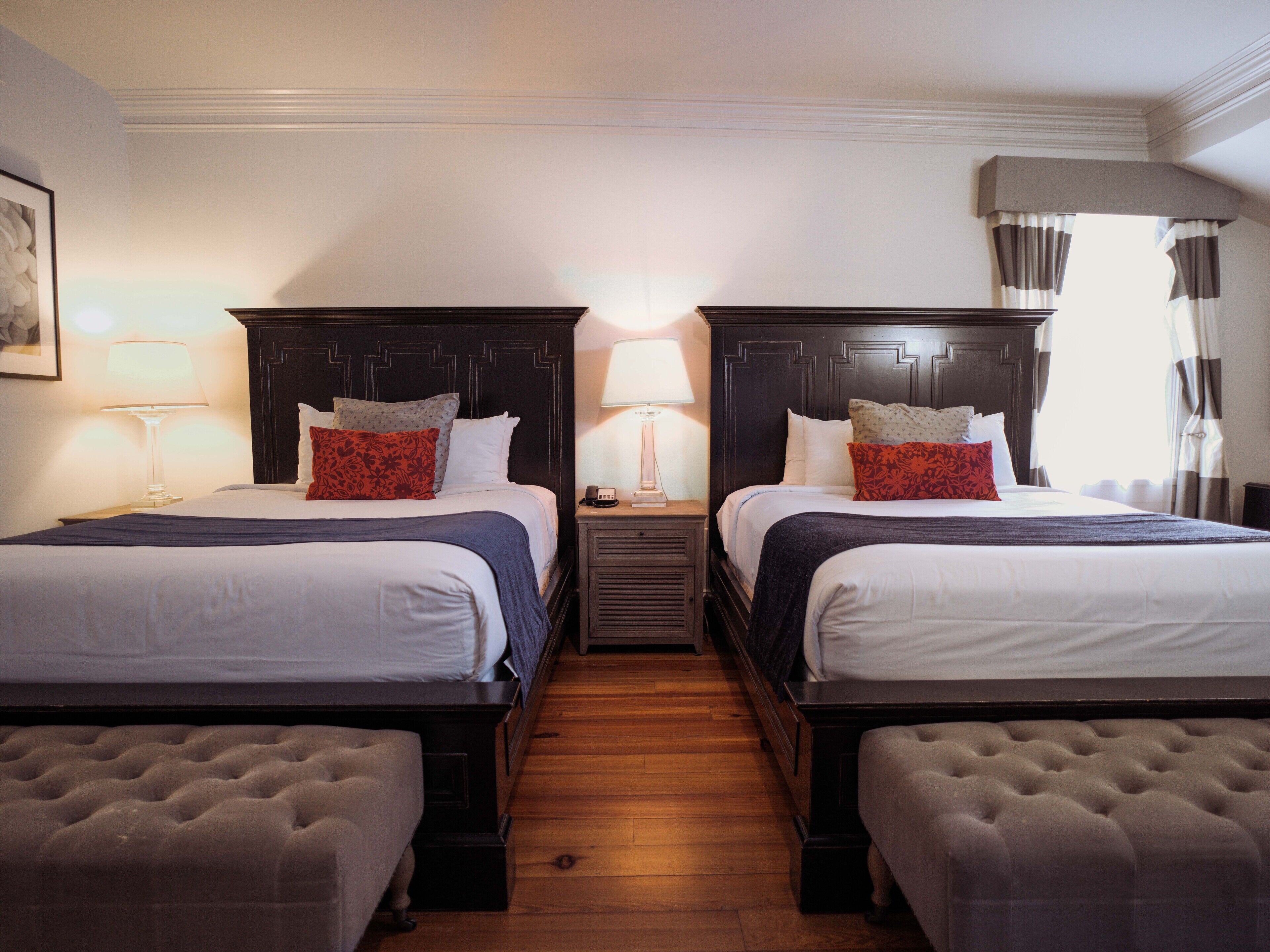 A hotel room with two beds, each with a headboard, bedside tables with lamps, and a wooden floor.