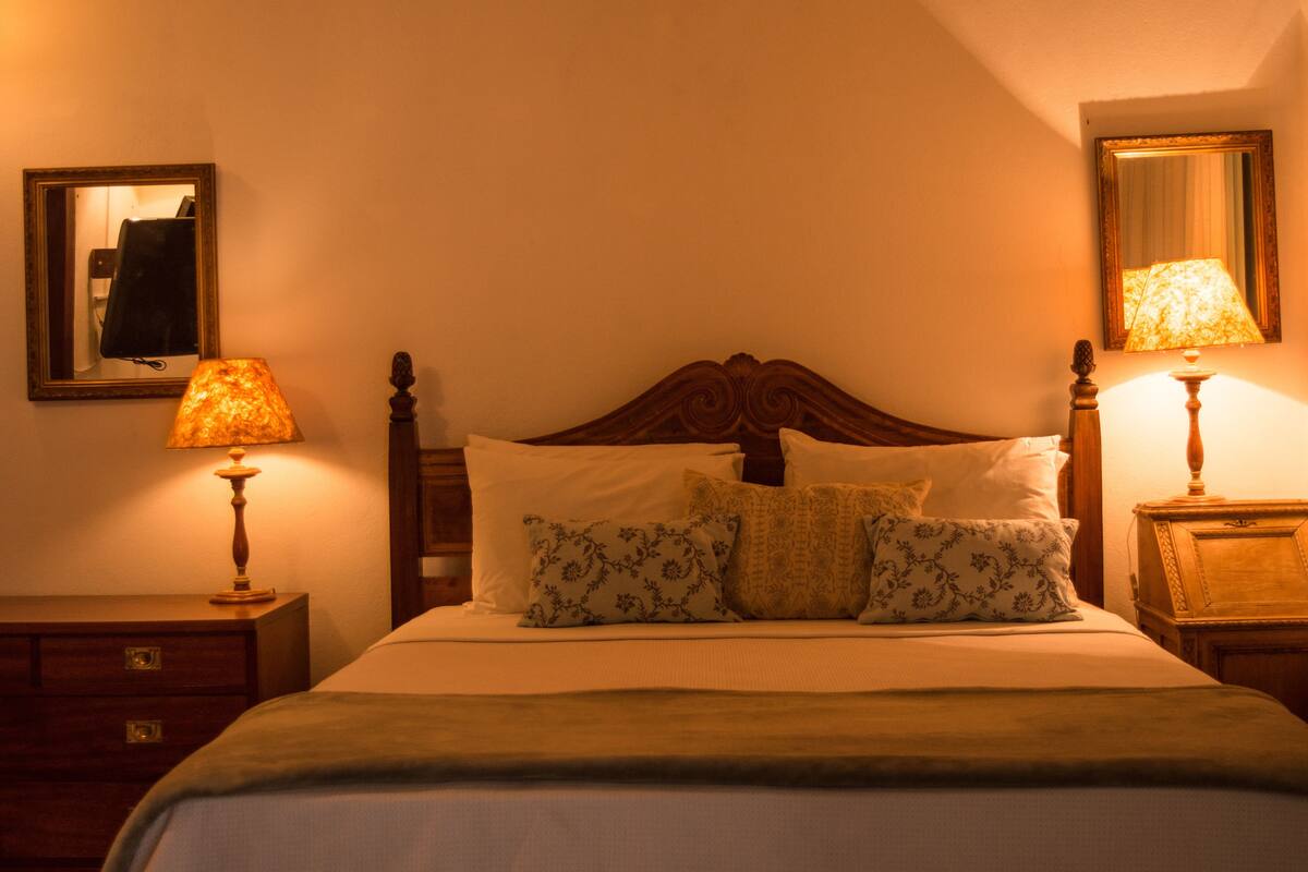 Un lit bien fait, agrémenté de coussins à motifs floraux, de deux lampes de chevet et d’un miroir au mur.
