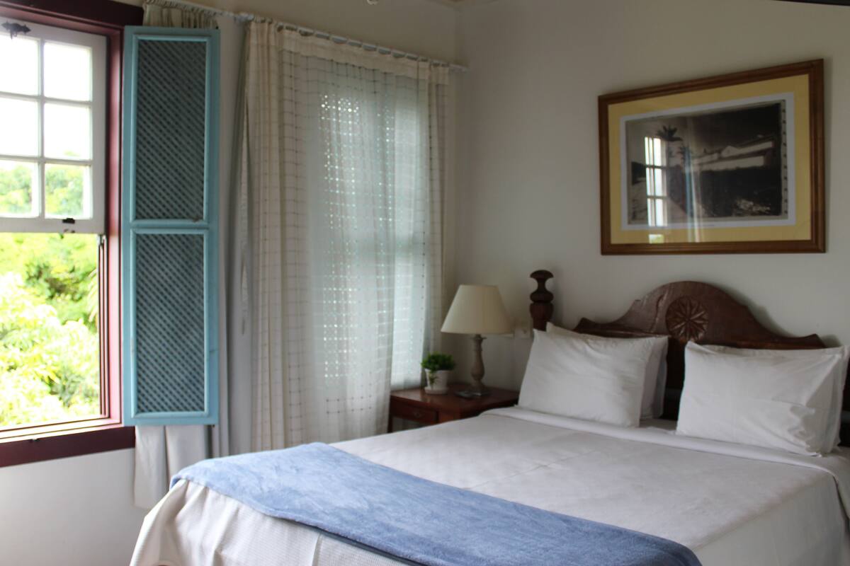 Un lit bien fait, recouvert d’un plaid bleu, une table de chevet avec une lampe, et une fenêtre avec des volets bleus.