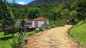 Un sendero de piedra conduce a un edificio blanco con techo de tejas rojas, rodeado de exuberante vegetación y una montaña al fondo.