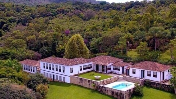 Una casa grande y blanca de dos pisos con piscina, rodeada de abundante vegetación y montañas al fondo.