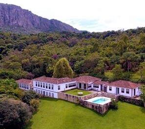 A large, white, two-story house with a swimming pool, surrounded by lush greenery and mountains in the background. - Caviúna Pouso Rural (Tiradentes)