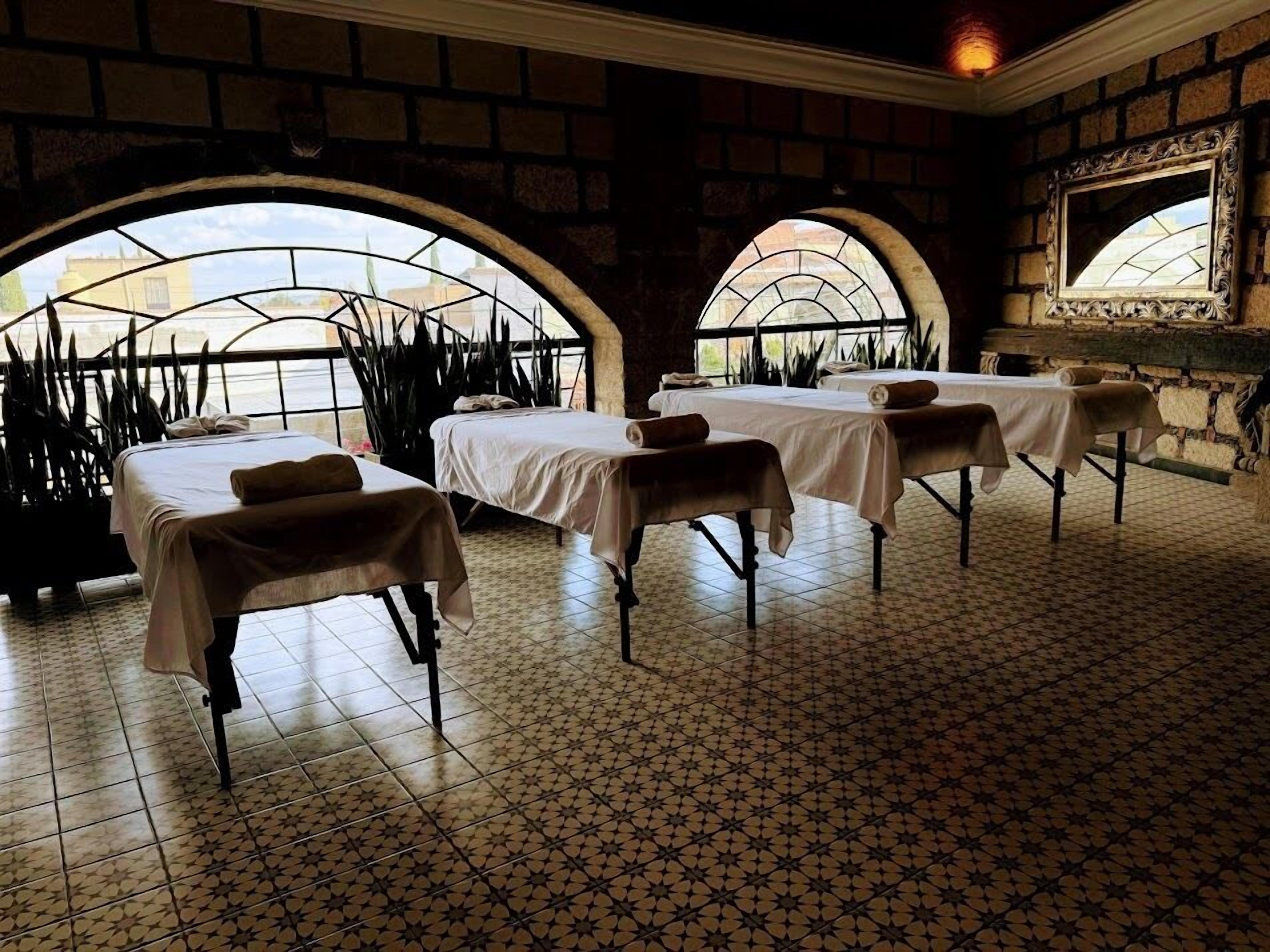 A spa room with three treatment tables, a brick wall, and large arched windows.