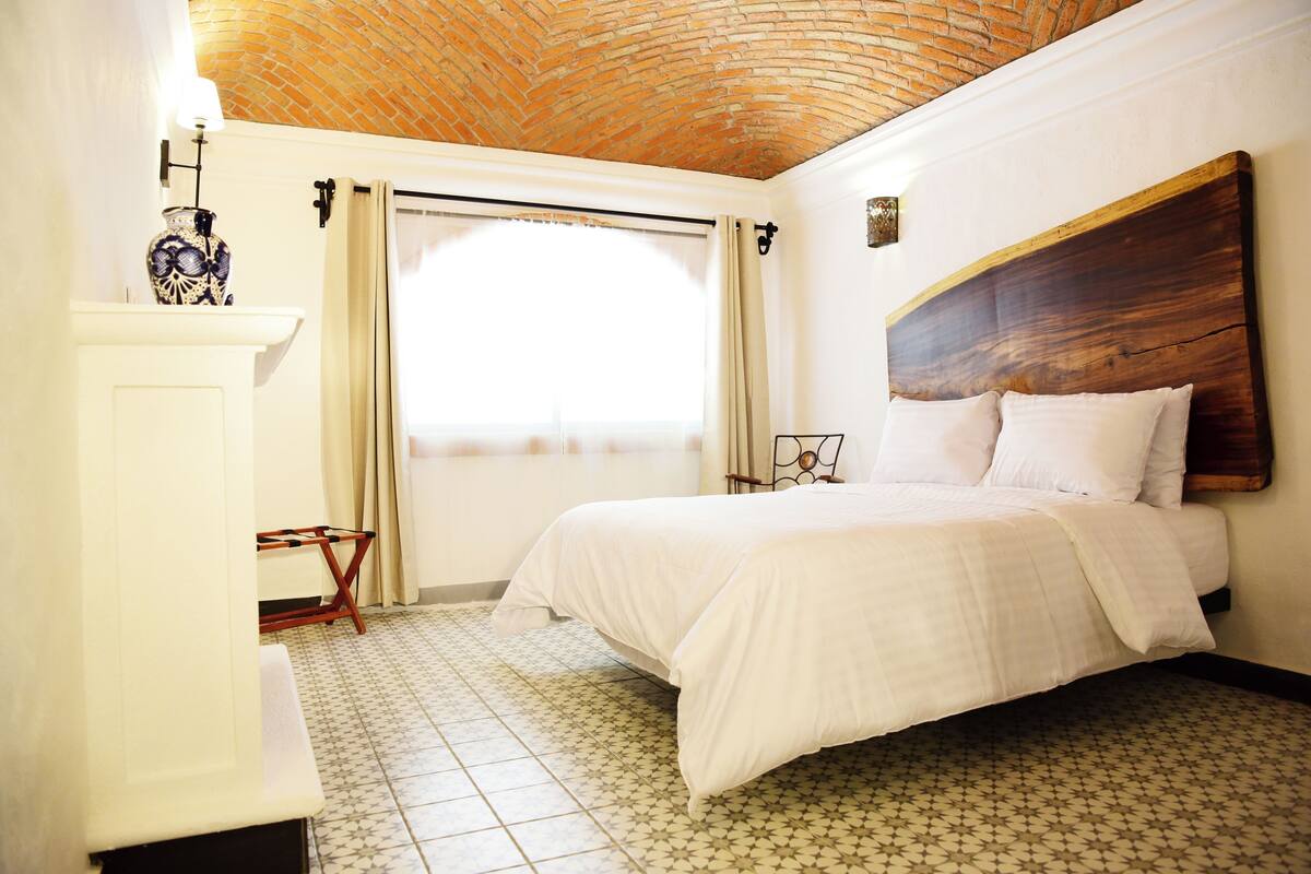A bedroom with a large bed, a wooden headboard, a tiled floor, a window with curtains, and a small table with a vase.