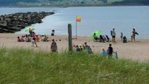 Una playa con gente relajándose y otros caminando por la orilla. Hay un asta con una bandera y algunos sombrillas.
